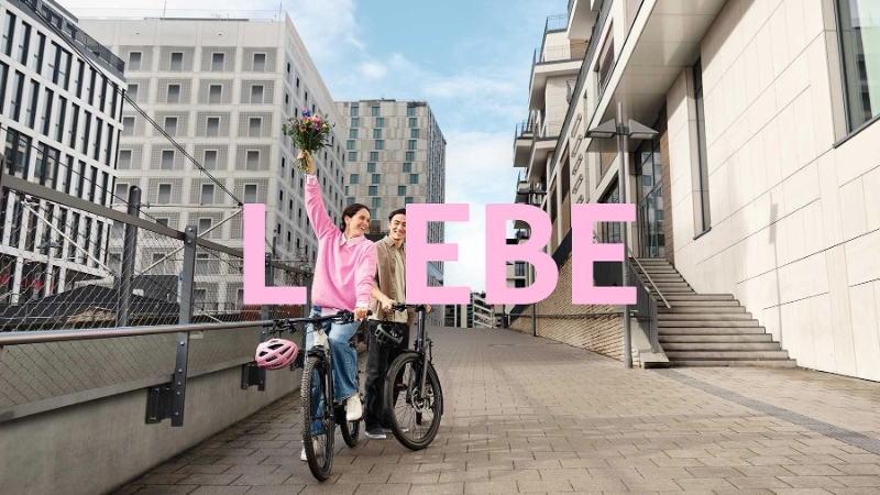 2 JobRadler:innen auf einem Fahrrad in einem urbanen Umfeld mit einem Blumenstrauß. 2 JobRadler:innen auf einem Fahrrad in einem urbanen Umfeld mit einem Blumenstrauß.